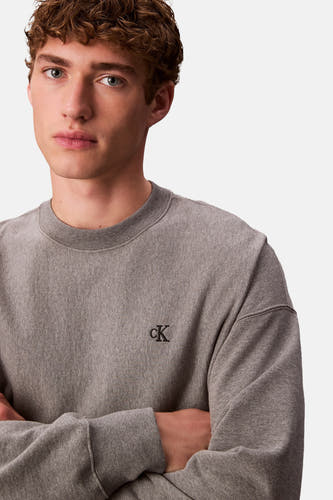 Pull-over gris de Calvin Klein, avec un petit logo noir sur la poitrine et des bras croisés.