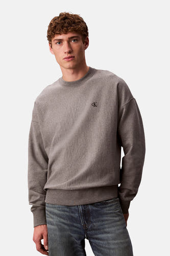 Pull-over gris de Calvin Klein, avec un petit logo 'ck' sur la poitrine, porté avec un jean bleu.