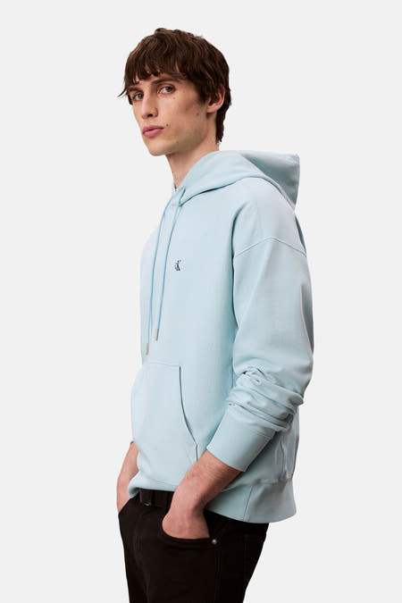 Pull-over bleu avec capuche de Calvin Klein, avec des cordons et une poche kangourou, porté avec un pantalon noir, en vue latérale.