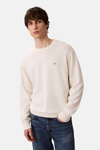 Gilet beige à col rond de Calvin Klein, avec un logo subtil sur la poitrine et porté avec des jeans.