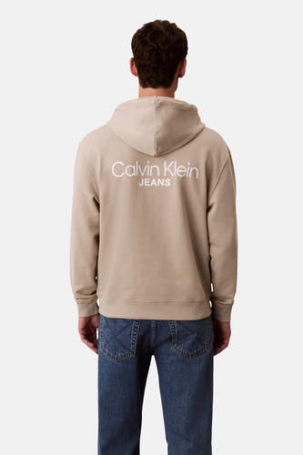 Beige sweater met kap van Calvin Klein Jeans, rugzijde met wit logo, gecombineerd met een donkerblauwe jeans.
