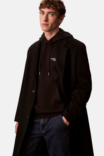 Pull-over noir de Calvin Klein avec capuche, porté sous une veste en laine noire et des jeans foncés.