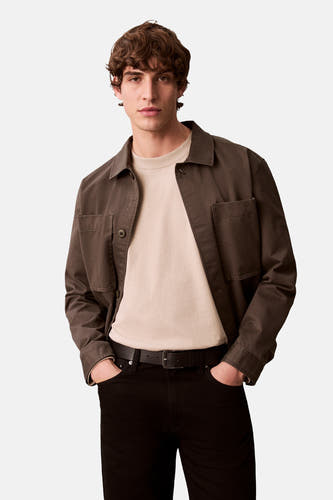 T-shirt beige à longues manches de Calvin Klein, porté sous un blouson marron avec fermeture à boutons et un pantalon noir avec ceinture.