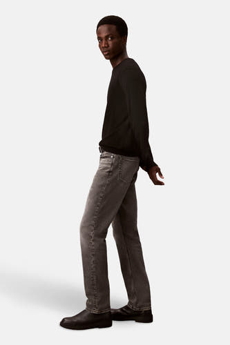 Man in zwarte trui, grijze jeans en zwarte schoenen, staand.
