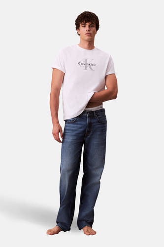 Jean droit en denim bleu de Calvin Klein, porté avec un t-shirt blanc et pieds nus.