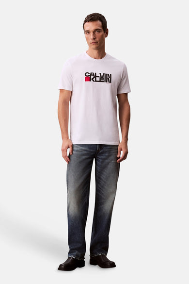 T-shirt blanc à manches courtes de Calvin Klein, avec un logo noir et rouge sur la poitrine, associé à des jeans et des chaussures noires.