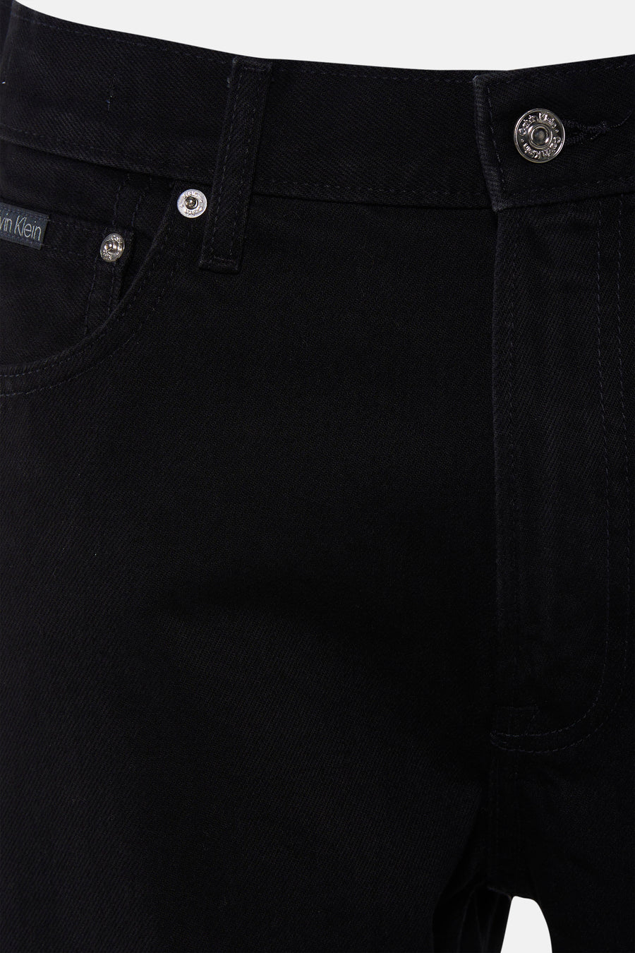 Jean droit noir de Calvin Klein, gros plan, avec boutons métalliques gravés du logo et petite étiquette en tissu sur la poche monnaie.