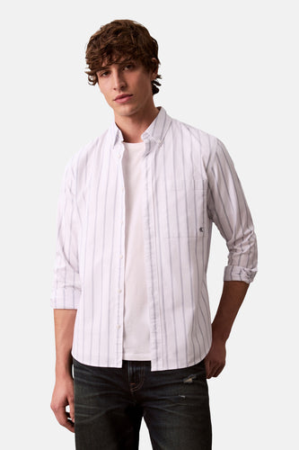 Man met bruin krullend haar, wit gestreept overhemd, wit T-shirt en donkere jeans.