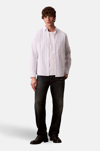 Chemise blanche à manches longues de Calvin Klein, portée sur un T-shirt blanc et associée à des jeans foncés et des chaussures noires.