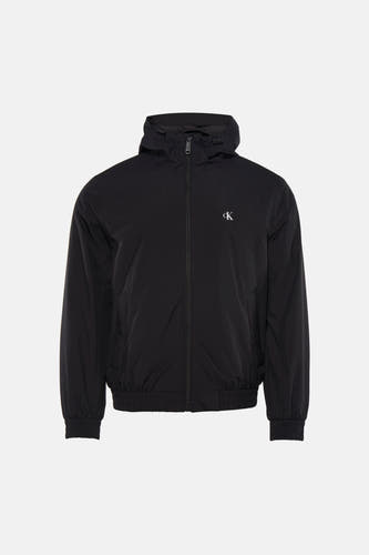 Manteau de pluie noir Calvin Klein avec capuche, porté sur une chemise noire et un jean gris, avec des baskets bleues.