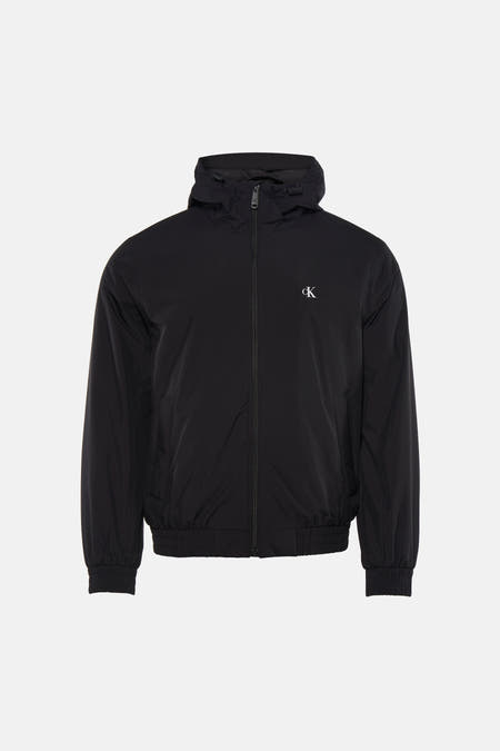 Manteau de pluie noir Calvin Klein avec capuche, porté sur une chemise noire et un jean gris, avec des baskets bleues.