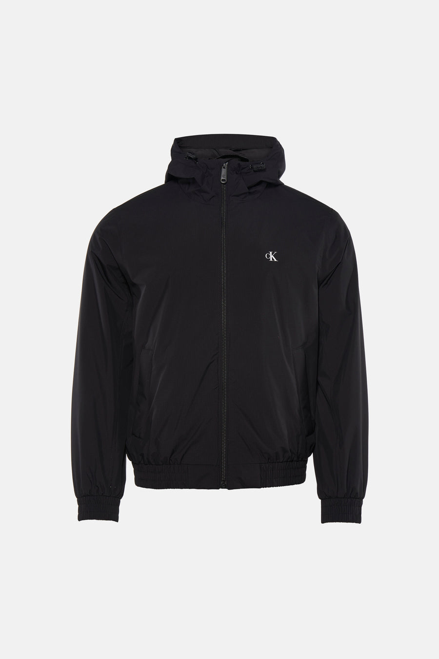 Manteau de pluie noir Calvin Klein avec capuche, porté sur une chemise noire et un jean gris, avec des baskets bleues.