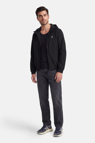 Zwarte regenjas van Calvin Klein met capuchon, gedragen over een zwart shirt en grijze jeans, met blauwe sneakers.
