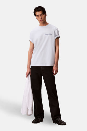 T-shirt bleu à manches courtes de Calvin Klein, porté avec un jean noir et des mocassins en cuir, avec une chemise blanche à la main.