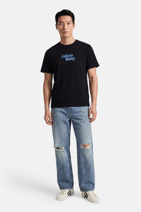 Zwart korte mouwen T-shirt met blauw logo van Calvin Klein, gedragen met gescheurde jeans en witte sneakers, front view