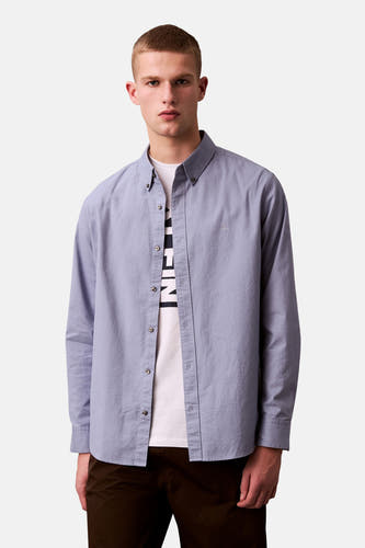 Chemise bleue à manches longues de Calvin Klein, portée sur un T-shirt blanc et un pantalon marron.