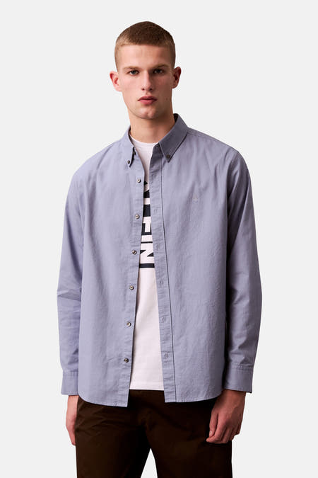 Chemise bleue à manches longues de Calvin Klein, portée sur un T-shirt blanc et un pantalon marron.