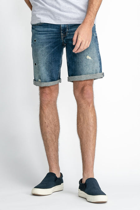 Jean en denim court de Petrol Industries, avec un pli et des déchirures, porté avec des baskets bleues et un T-shirt gris, vue latérale.