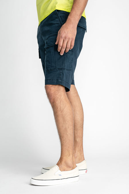 Short cargo bleu de Petrol Industries®, profil latéral, avec grandes poches et ourlet retroussé, porté avec un haut jaune et des baskets blanches