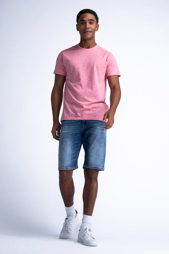Een man draagt een roze T-shirt en blauwe spijkerbroekshorts, met witte sokken en sportschoenen op een witte achtergrond.