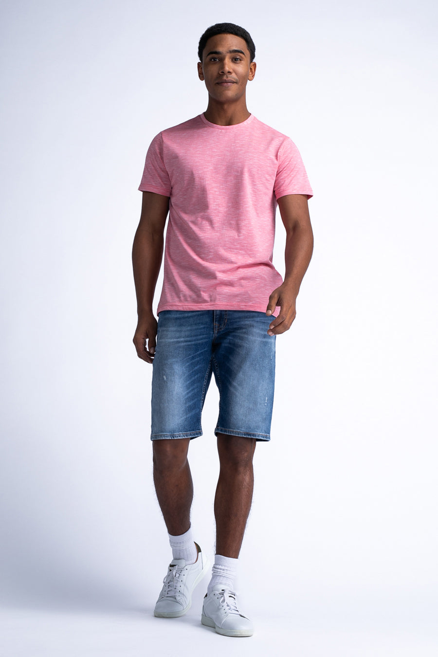 Een man draagt een roze T-shirt en blauwe spijkerbroekshorts, met witte sokken en sportschoenen op een witte achtergrond.