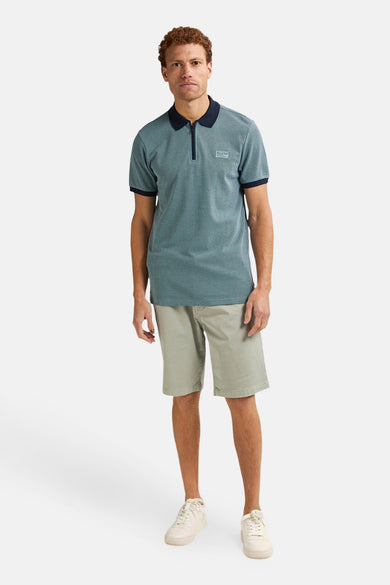 Man in blauwgroene polo en kaki shorts, staand op een witte achtergrond.