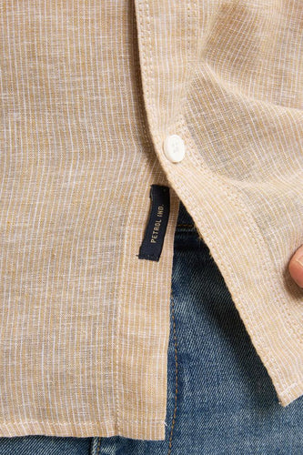 Beige linnen overhemd met knoop en donkerblauw merklabel, gedragen over een blauwe spijkerbroek.