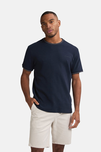 T-shirt bleu à manches courtes de Petrol Industries, avec un col et des manches bleu clair, porté avec des shorties beiges.