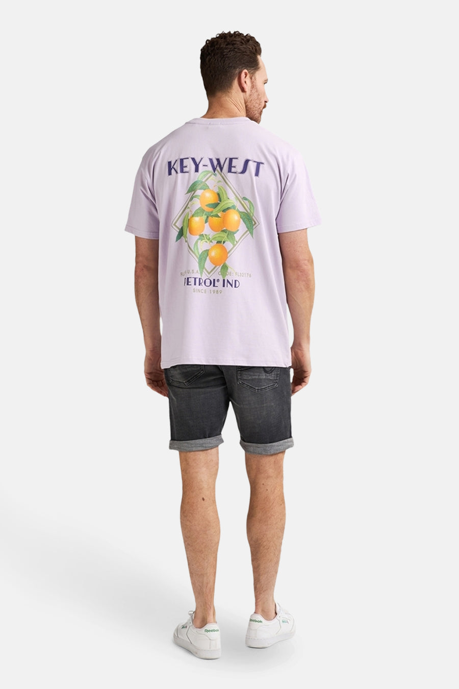 Een man van achteren gezien in een lila T-shirt met sinaasappeldruk, zwarte denim shorts en witte sneakers.
