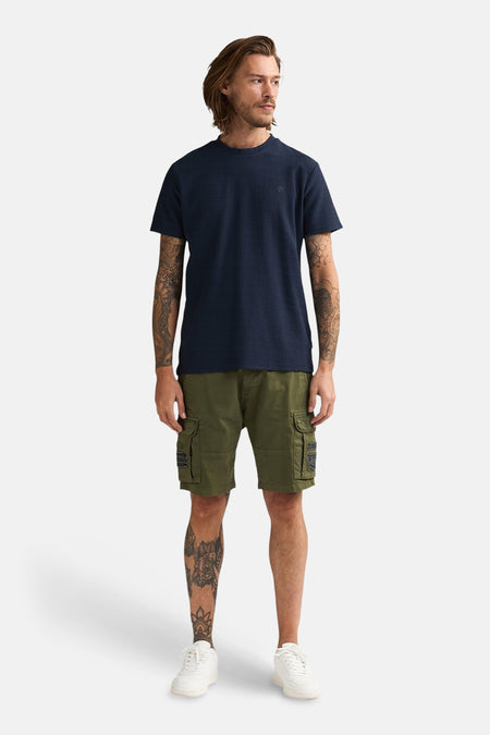 T-shirt bleu à manches courtes de Petrol Industries®, avec une texture subtile et porté avec un short kaki et des baskets blanches.