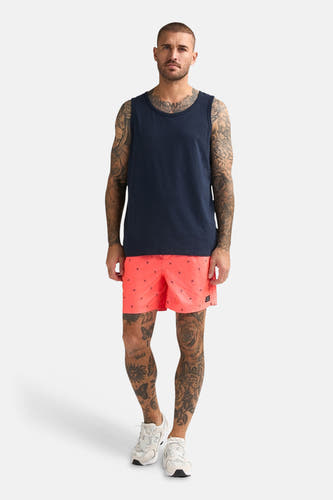 Débardeur bleu de Petrol Industries, porté par un homme tatoué avec un short rouge corail à motif d'ancre et des baskets blanches.