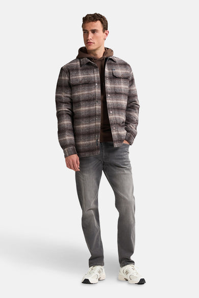 Veste courte brune de Petrol Industries, avec un motif à carreaux et une hoodie brune en dessous, associée à des jeans gris et des baskets blanches.