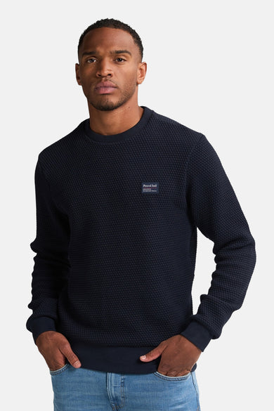Pull-over bleu de Petrol Industries avec une structure subtile en losange et un col rond, porté avec un pantalon de cuir.
