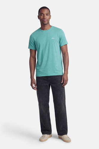 Groen T-shirt met korte mouwen van Guess®, met kleine tekstprint op de borst, gecombineerd met donkere jeans en beige sneakers.