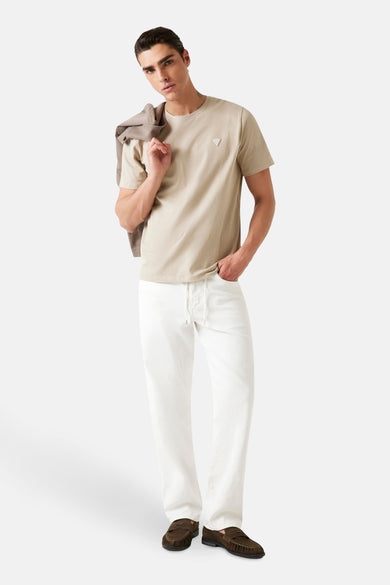 T-shirt beige à manches courtes de Guess, porté avec un manteau sur l'épaule et un pantalon blanc.