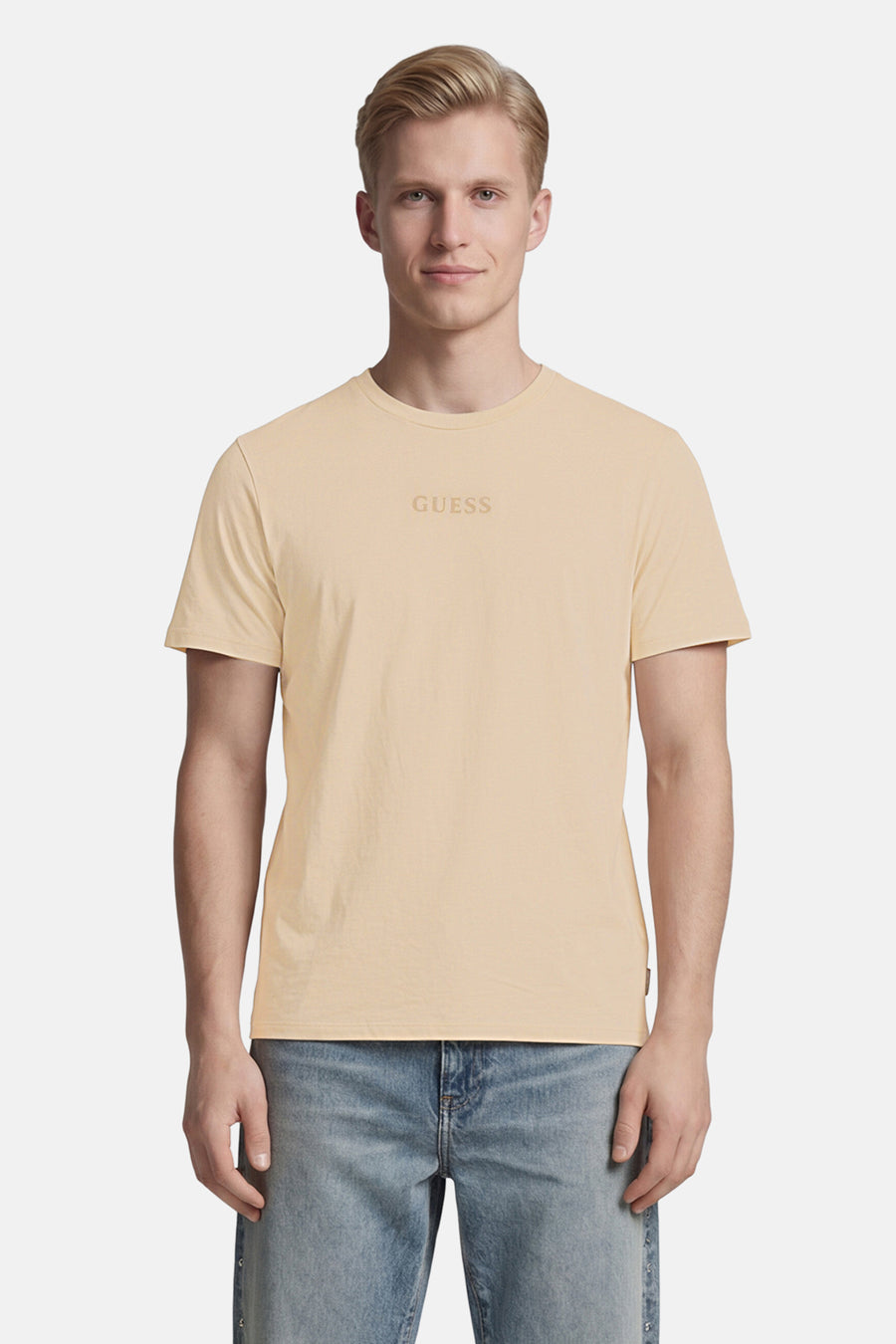T-shirt beige de Guess avec logo GUESS ton sur ton sur la poitrine, porté par un homme avec un jean bleu clair.