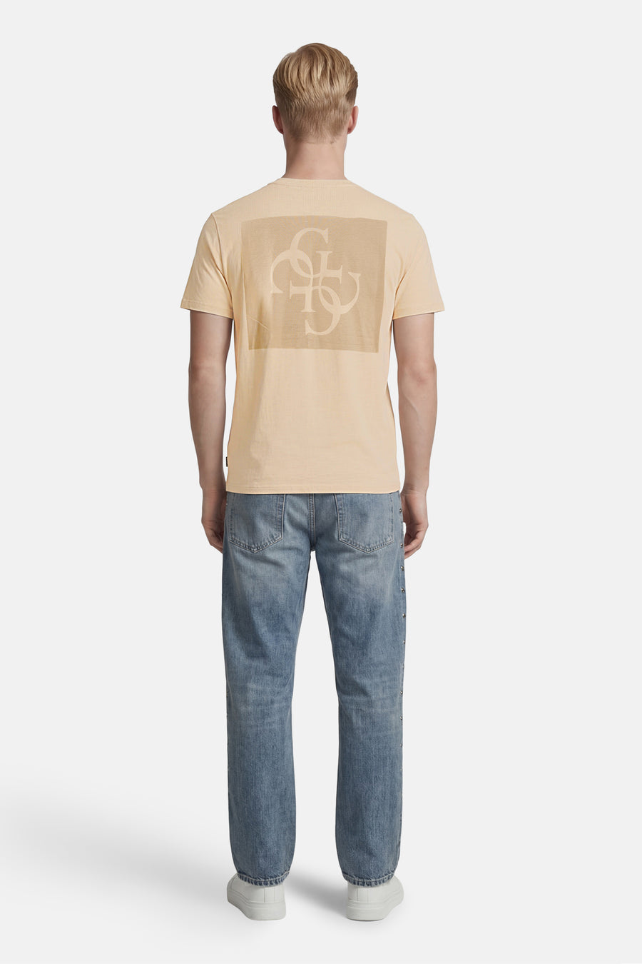 T-shirt beige Guess avec logo ton sur ton au dos, porté par un mannequin avec un jean clair clouté et des baskets blanches, vue arrière.
