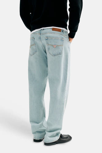 Jean en denim bleu clair droit de Guess, avec patch en cuir marron et logo triangulaire sur la poche arrière droite, porté avec des mocassins noirs, vue arrière.