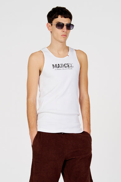 Débardeur blanc de Le Fabuleux Marcel de Bruxelles, avec un imprimé logo et porté avec un pantalon marron.