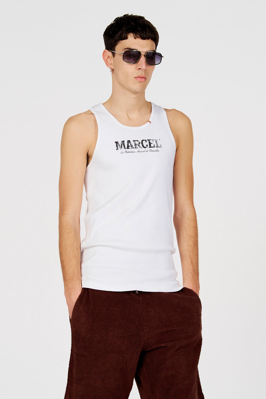 Débardeur blanc de Le Fabuleux Marcel de Bruxelles, avec un imprimé logo et porté avec un pantalon marron.