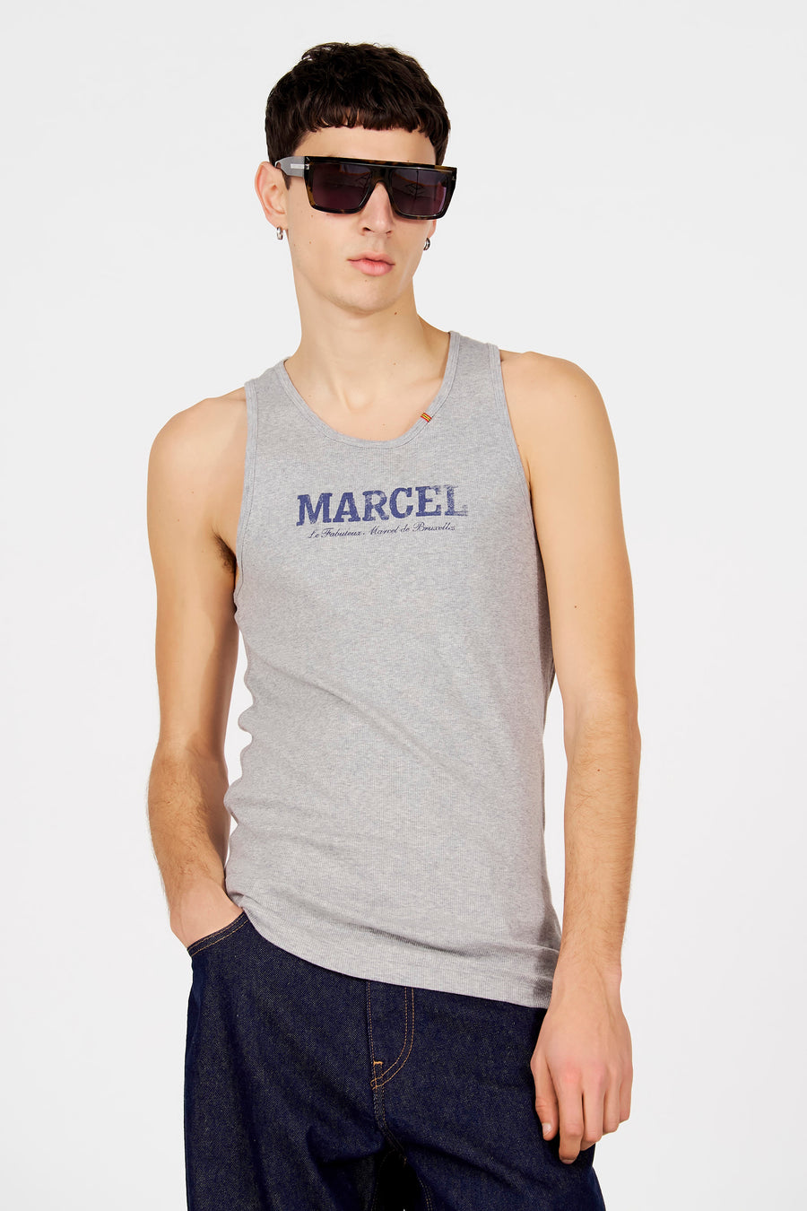 Grijs tanktop van Le Fabuleux Marcel de Bruxelles, met blauwe tekstprint op de borst, gedragen met een donkere jeans en zonnebril.
