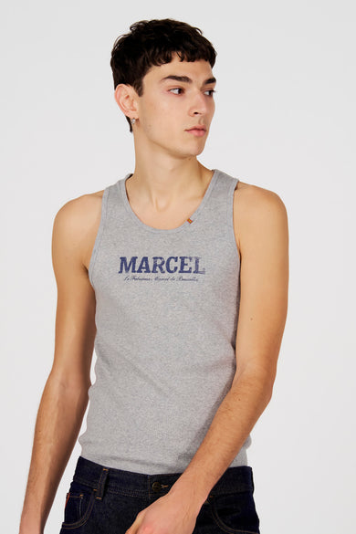 Débardeur gris de Le Fabuleux Marcel de Bruxelles, avec imprimé texte bleu sur le devant, porté avec des lunettes de soleil et une boucle d'oreille argentée.