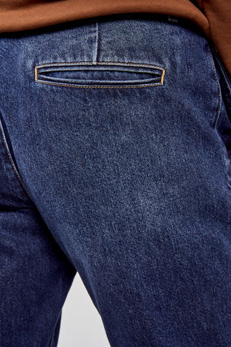 7/8 jeans en denim bleu moyen de Le Fabuleux Marcel de Bruxelles, avec un sac à dos et une chemise brune.