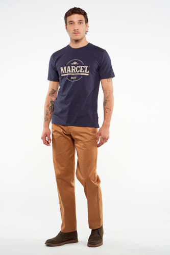 Pantalon de costume brun du Le Fabuleux Marcel de Bruxelles, porté avec un T-shirt bleu marine et des bottes kaki.