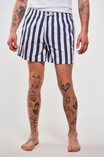 Pantalon de bain rayé bleu de Le Fabuleux Marcel de Bruxelles, avec des tatouages sur les jambes et les bras.