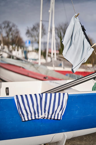 Pantalon bleu de Le Fabuleux Marcel de Bruxelles, rayé, suspendu à une corde à linge à côté d'un bateau.