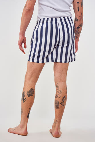 Pantalon bleu de Le Fabuleux Marcel de Bruxelles avec des rayures verticales, porté avec un t-shirt blanc et des tatouages.