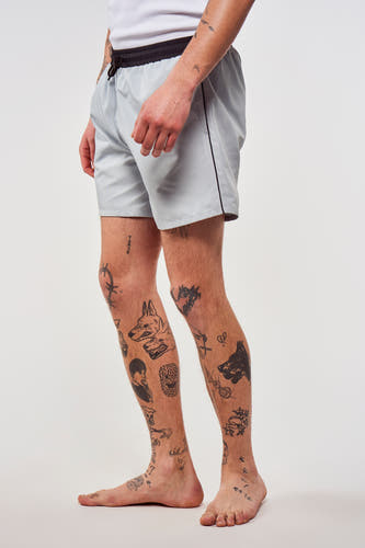 Pantalon bleu de Le Fabuleux Marcel de Bruxelles, avec une ceinture noire et des tatouages sur les jambes, en vue latérale.