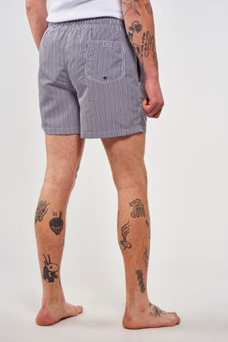 Pantalon de bain rayé bleu de Le Fabuleux Marcel de Bruxelles, avec des tatouages sur les jambes.