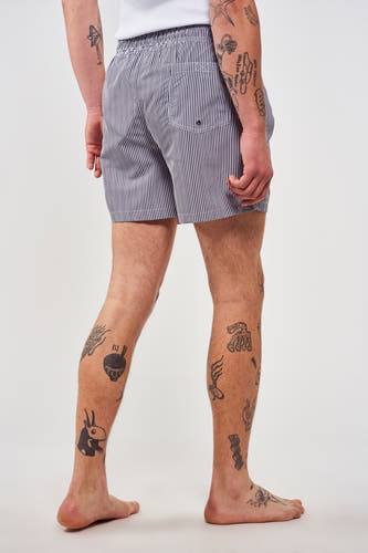 Pantalon de bain rayé bleu par Le Fabuleux Marcel de Bruxelles, avec une ceinture élastique et des tatouages sur les jambes.